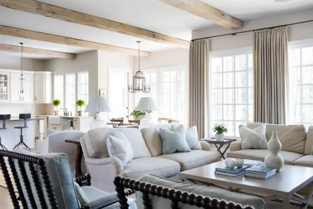Living room with neutral-toned sofas, light blue accent pillows, and dark wood chairs. Exposed wooden ceiling beams and large windows with beige curtains are visible.