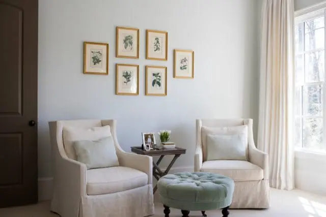 Light blue walls with a gallery of six framed botanical prints, two slipcovered armchairs with light blue pillows, and a tufted teal ottoman.