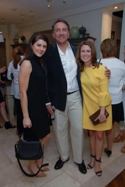 Man in a dark blazer and white shirt stands between two women, one in a black sleeveless dress and the other in a yellow dress with bell sleeves.