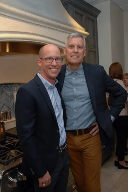 Two men smiling, one with glasses and a dark suit, the other in a blue shirt and dark suit. They are standing in front of a kitchen range and cabinetry.