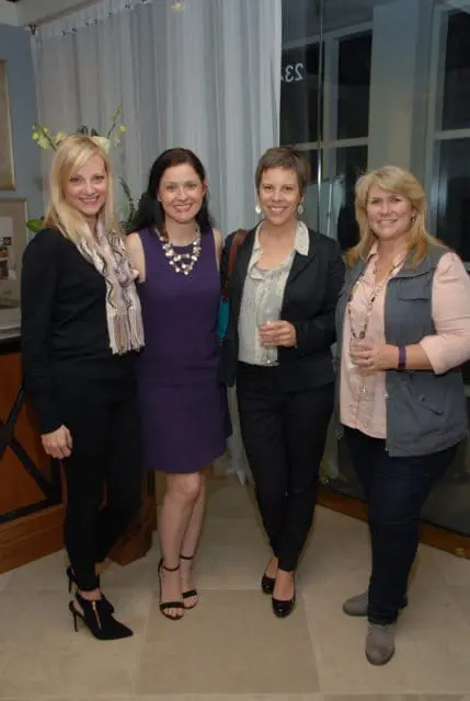 Four women smiling, two holding glasses, in a well-lit indoor setting with sheer curtains.