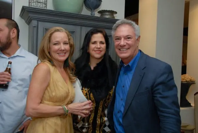 Three people smiling at the camera: a woman in a gold dress, a woman in a black and gold patterned jacket with a fur collar, and a man in a blue shirt and navy blazer.
