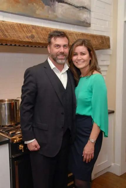 Man in a dark suit and woman in a green top and navy skirt pose in a kitchen with a large wooden beam above a white brick backsplash.
