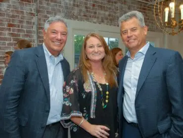 Three people smiling at the camera in front of a brick wall. The two men are wearing suits, and the woman is wearing a floral top and a long necklace.
