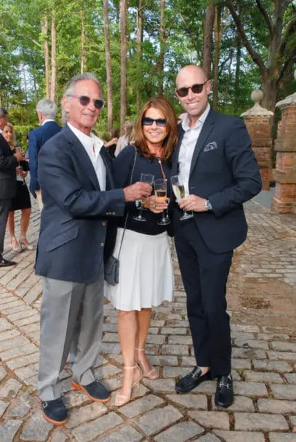 Three people in formal attire holding champagne glasses outdoors, with trees and a brick structure in the background.