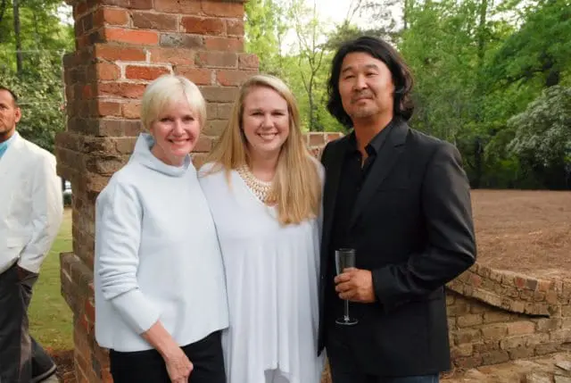 Three people posing in front of a brick structure and trees. A woman in a light blue top and black pants, a woman in a white top with a statement necklace, and a man in a black suit holding a champagne flute.