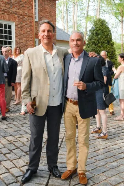 Two men stand with arms around each other's shoulders on a cobblestone patio. The man on the left wears a tan blazer over a light-colored shirt and dark pants, holding a beer bottle. The man on the right wears a navy blazer over a patterned shirt and tan pants, holding a champagne flute.