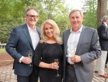 Three people in formal attire holding champagne flutes outdoors. A man in a dark suit and glasses stands on the left, a woman in a sparkly black dress in the center, and a man in a gray suit on the right.