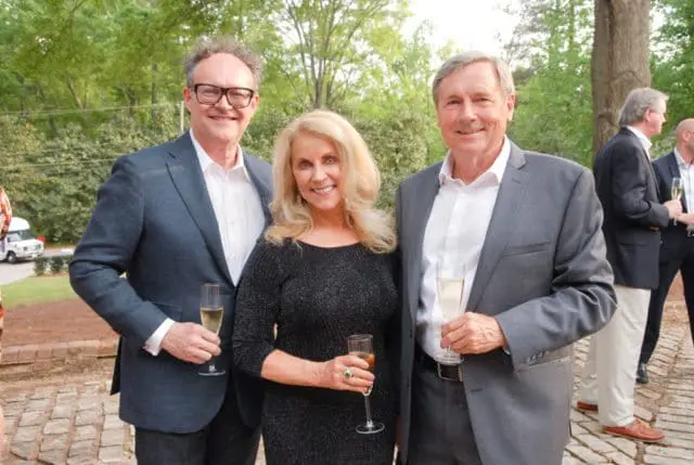 Three people in formal attire holding champagne flutes outdoors. A man in a dark suit and glasses stands on the left, a woman in a sparkly black dress in the center, and a man in a gray suit on the right.