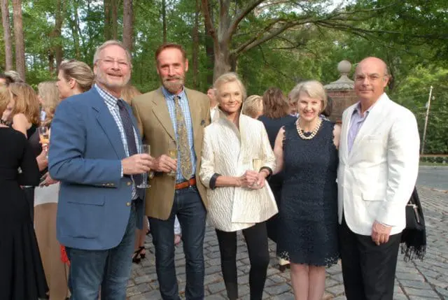 Five people posing for a photo outdoors, holding champagne flutes. A man in a blue blazer stands left of center, next to a man in a tan jacket. A woman in a cream jacket is next, followed by a woman in a navy dress, and a man in a white blazer on the right.