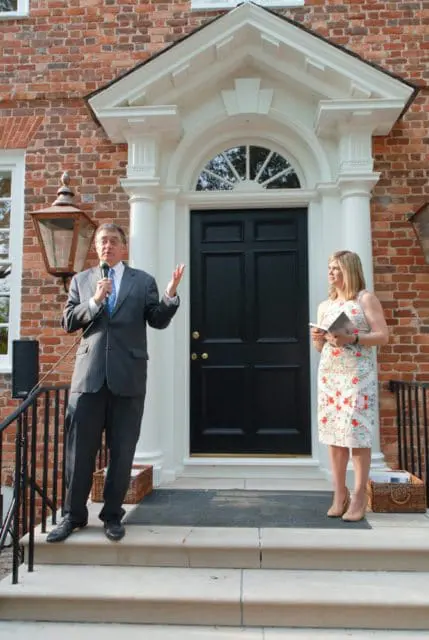 Man in suit speaking into microphone on steps of brick house with black front door and white arched entryway. Woman in floral dress stands nearby.