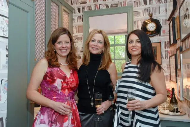 Three women smiling and holding champagne flutes in a room with patterned wallpaper and decorative clocks on the wall.