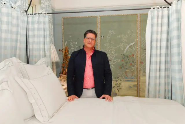 Man in a pink shirt and dark jacket standing behind a bed with white linens. Behind him is a screen with a bird and floral motif and light blue checkered curtains.