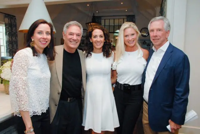 Five people posing for a photo indoors. A woman in a white lace top stands next to a man in a tan jacket. A woman in a white dress is in the center, flanked by a woman in a white ruffled top and a man in a navy blazer.