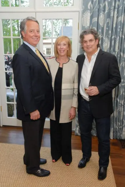 Three people pose for a photo indoors. A man in a dark suit and yellow tie stands on the left. A woman in a patterned jacket and black pants stands in the middle. A man in a dark jacket and jeans holds a glass of wine on the right.