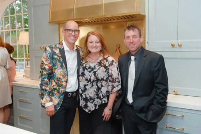 Three people posing in a kitchen with light blue cabinetry, gold hardware, and a gold range hood. The person on the left wears a colorful floral blazer.