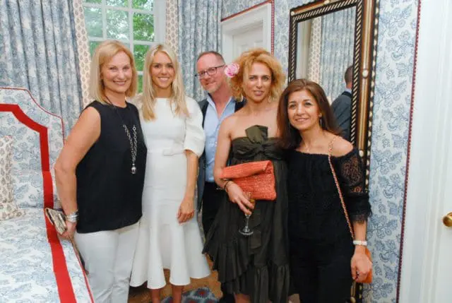 Five people pose for a photo in a room with blue and white patterned wallpaper and curtains. A woman in black stands left, next to a woman in white. Behind them are three people, two women and one man.