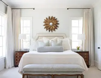 Serene bedroom with a king-size bed dressed in white linens, flanked by two nightstands with lamps. A sunburst mirror hangs above the headboard, and floor-to-ceiling curtains frame the windows.