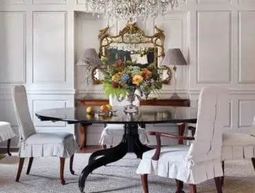 Formal dining room with a dark oval table, upholstered chairs with white slipcovers, a crystal chandelier, and a gilded mirror above a floral arrangement.