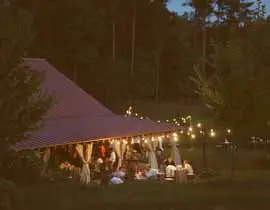 Open-air pavilion with a red metal roof, draped curtains, and string lights, set in a wooded area at dusk.