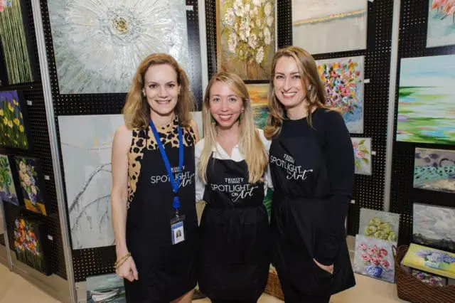Three women smiling in front of a display of artwork. They are wearing black aprons with "Trinity School Spotlight on Art" printed on them.