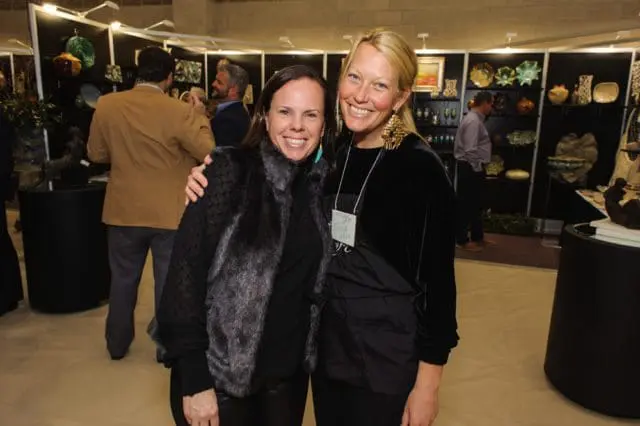 Kelly Buschmann and Tricia Collie smile at the camera, standing in front of displays of art and pottery at an event.