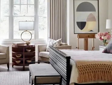 Bedroom with a dark wood bed frame, plush gray pillows, and a tan throw blanket. A light-colored armchair sits near a round wooden side table with a lamp.
