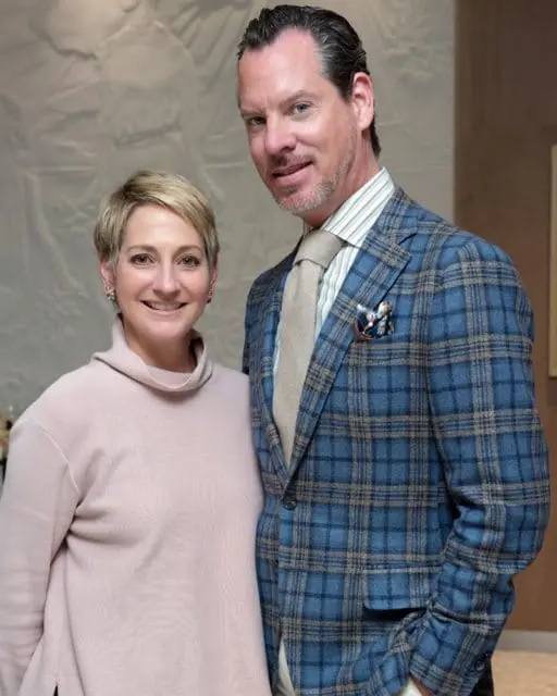 A woman with short blonde hair wearing a pink turtleneck sweater stands next to a man with dark hair wearing a blue plaid suit jacket, a beige tie, and a striped shirt.