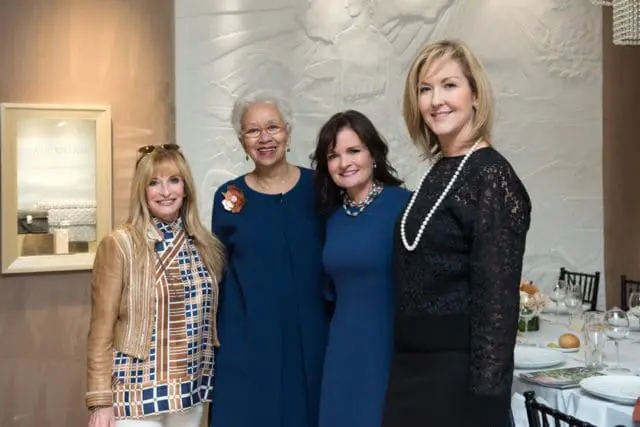 Four women pose for a photo in front of a textured wall. A framed display with the word VALENTINO is visible to the left.