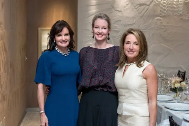 Three women pose for a photo in a dining room. The woman on the left wears a blue dress and pearl necklace. The woman in the middle wears a dark, patterned top and black skirt. The woman on the right wears a cream-colored sleeveless top and pearl necklace.