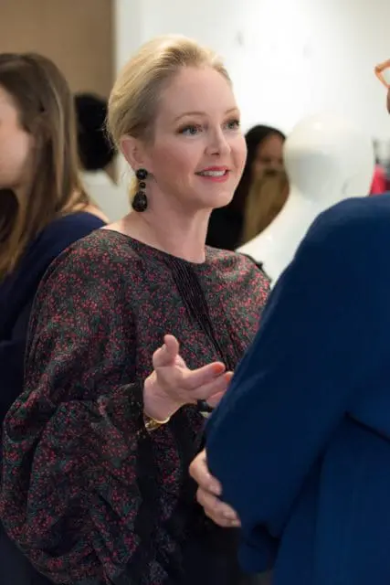 Blonde woman with dark earrings and a floral print blouse speaking to someone in a blue jacket.