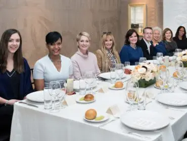 A group of eight people are seated at a long, white-clothed table set for a meal. Floral centerpieces adorn the table.