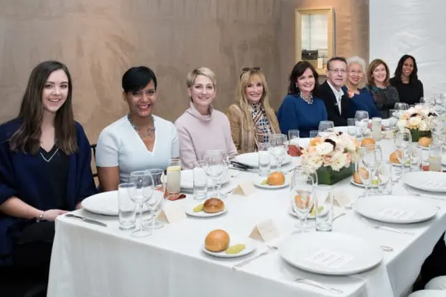 A group of eight people are seated at a long, white-clothed table set for a meal. Floral centerpieces adorn the table.