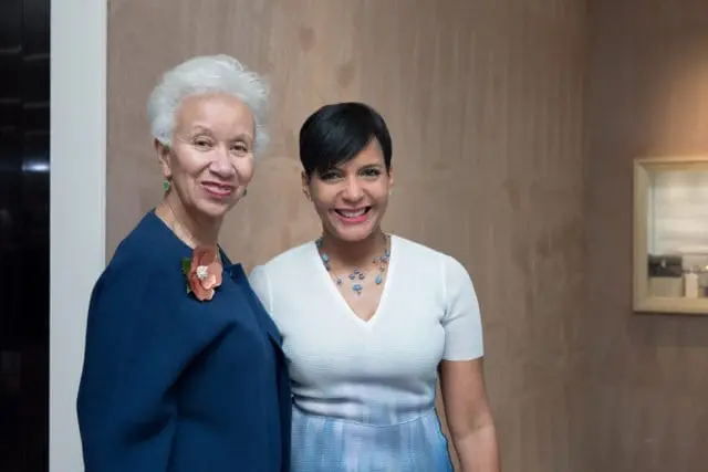 Two women smiling at the camera. The woman on the left has white hair and wears a navy blue jacket with a floral brooch and green earrings. The woman on the right has short black hair and wears a white top and a blue and white patterned skirt.