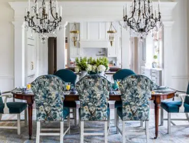 Elegant dining room with a dark wood table surrounded by upholstered chairs in teal velvet and a blue floral pattern. Two ornate crystal chandeliers hang above, and a glimpse of a white kitchen is visible in the background.