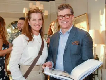 Woman in a white shirt and tan skirt and man in a blue suit jacket and glasses holding an open book