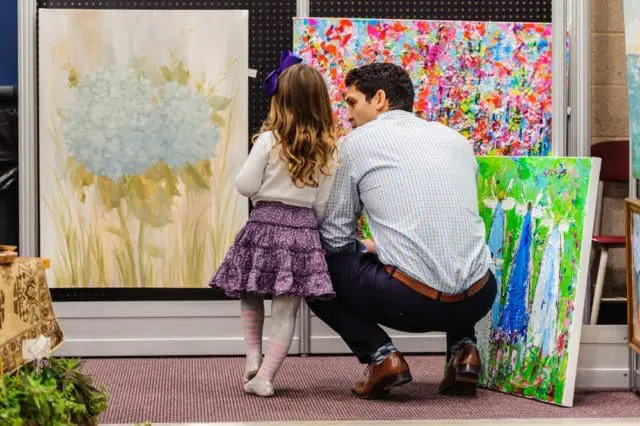 Man and young girl look at paintings at an art market; one painting is of a blue hydrangea, another is abstract floral, and a third depicts figures in blue dresses.