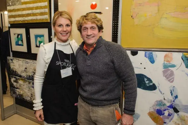 A woman wearing a black apron with 'Spotlight Art' and 'Trini School' visible, stands next to a man in a grey sweater and tan pants. They are posing in front of abstract paintings.