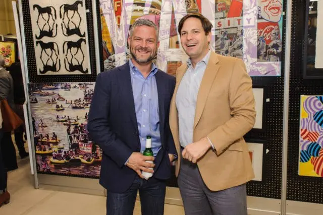 Two men smiling, one in a navy blazer holding a beer bottle, the other in a tan blazer, with colorful artwork behind them.
