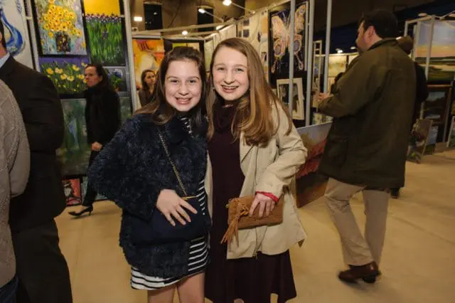 Two young women smiling in front of art displays at an exhibition. One wears a dark fuzzy coat over a striped dress, the other a tan trench coat over a burgundy dress.