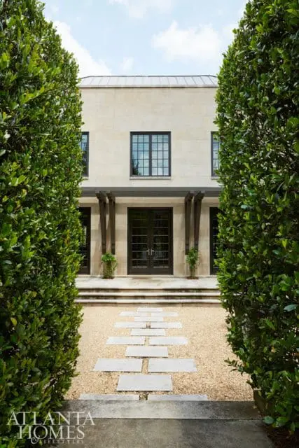 Stone facade house with dark-paned windows, double glass doors, and a stone pathway through a gravel garden bordered by manicured hedges.