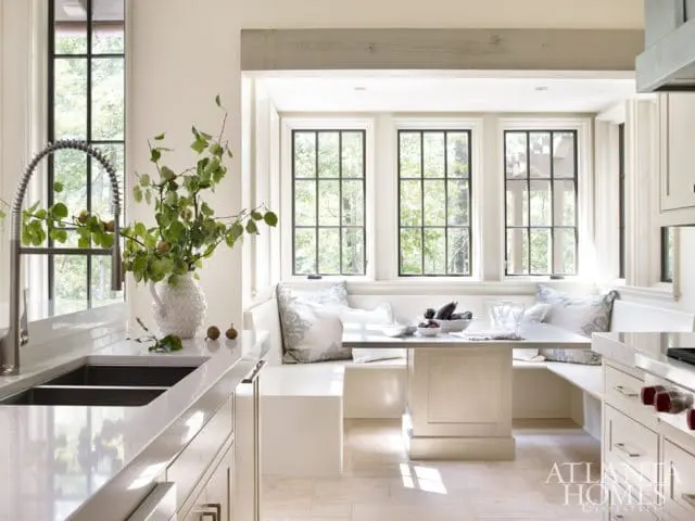 Cream kitchen with built-in banquette seating, white quartz countertops, double sink, and a table set with fruit. Large windows overlook trees.