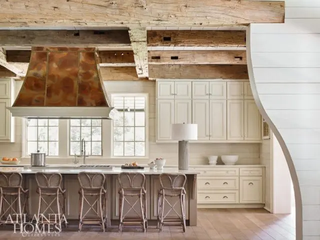 Cream kitchen with a copper range hood, exposed wooden beams, and a large island with wicker bar stools.