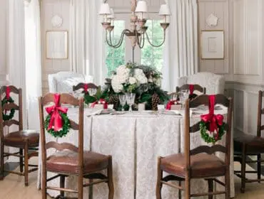 Dining room with a table set for a holiday meal, featuring chairs adorned with wreaths and red bows, a floral centerpiece, and paneled walls.