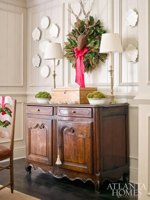 Dark wood antique buffet with ornate carvings, topped with two lamps, a wooden box, and moss bowls. A magnolia wreath with a deer head and red ribbon hangs on the paneled wall above.