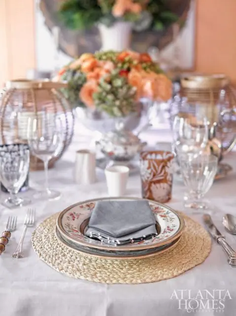 Table setting with floral plates, gray napkins, woven placemats, crystal glassware, and a centerpiece of peach and green flowers.