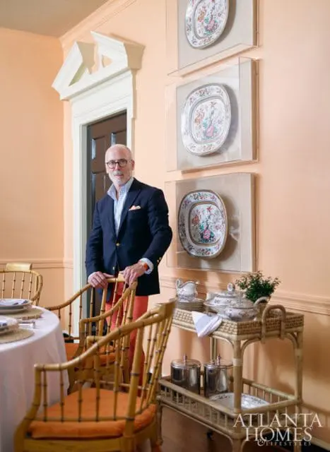 Man in navy blazer and coral pants stands by bamboo chairs in a room with peach walls, three framed decorative plates, and a wicker bar cart with serving dishes.