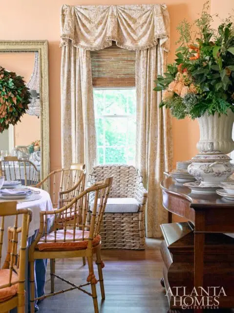 Dining room with peach walls, bamboo chairs with orange cushions, a wicker chair, and a large floral arrangement on a wooden table.