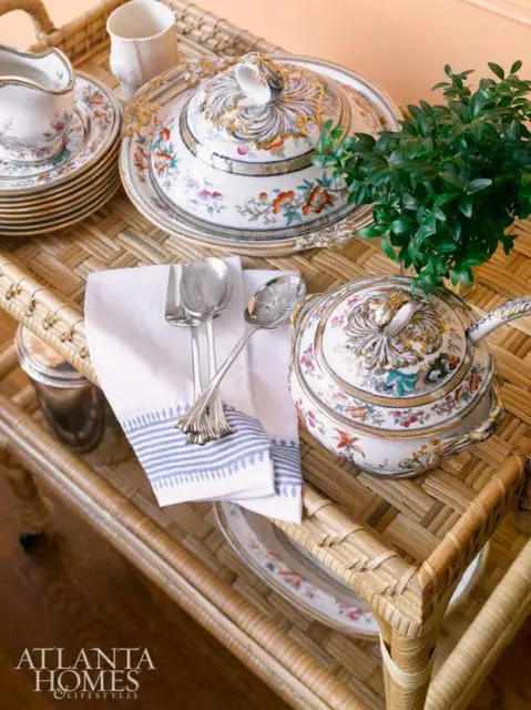 White ceramic serving dishes with ornate floral patterns and gold trim, a stack of plates, silverware, and a green plant on a woven wicker tray.