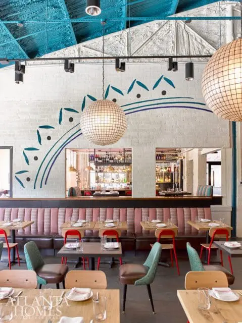 Restaurant dining area with pink and brown tufted banquettes, wooden tables, red chairs, and large disco ball light fixtures. A mural of teal arches and dots adorns the white brick wall.